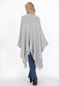 Harmaa neulotun poncho, jossa on fringe-reunat ja löysä muotoilu, joka laskeutuu kehon päälle. Yhdistetty sinisiin farkkuihin ja mustiin korokepolkimiin.