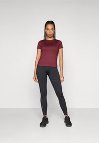 Bordeauxrotes T-Shirt mit kurzen Ärmeln, kombiniert mit schwarzen Leggings mit subtiler Textur. Das Model steht in schwarzen Sportschuhen vor einem neutralen Hintergrund.
