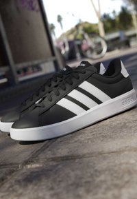 Baskets Adidas noires avec semelle blanche et trois bandes blanches sur le côté, reposant sur une surface extérieure pavée près d'un vélo en arrière-plan.