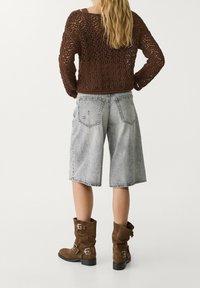 Suéter marrón de ganchillo, pantalones cortos de mezclilla gris con bordes deshilachados, y botas de ante marrón hasta la mitad de la pantorrilla con hebillas. Las botas tienen un acabado texturizado.