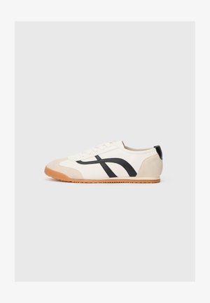 Krém színű vászon sportcipő, fekete bőr díszítésekkel, texturált gumitalppal és ívelt fekete mintázattal az oldalán.