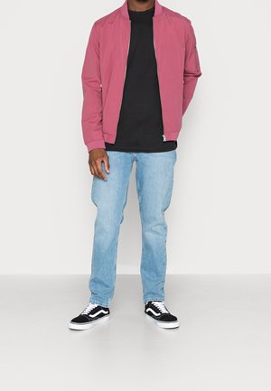 Homme portant une veste bomber rose sur un t-shirt noir, un jean bleu clair et des baskets noires et blanches, debout avec une main derrière le dos.