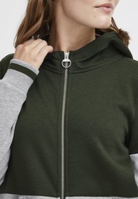 Grön zip-up hoodie med hög krage, grå ärmar och silverblixtlås. Mjuk tygkvalitet med en slät textur och kontrasterande färgdetaljer.
