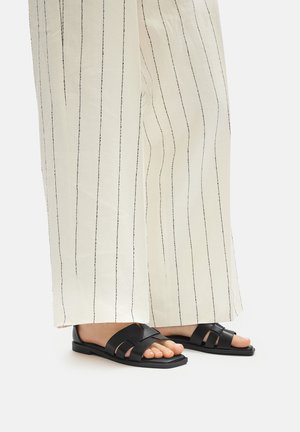 Sandale din piele neagră cu talpă plată și design cu curea împletită, purtate cu pantaloni largi cu dungi crema, evidențiind degetele de la picioare.