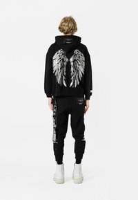 Sudadera negra con un diseño de alas de ángel plateadas en la espalda, combinada con joggers negros que presentan detalles gráficos y botas blancas hasta el tobillo.