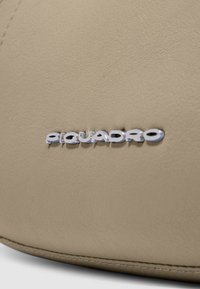 Superficie in pelle beige con trama fine; presenta un logo "Piquadro" metallico argentato; cuciture visibili lungo il bordo.