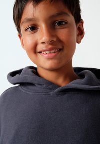 Garçon souriant au teint moyen portant un sweat à capuche gris foncé, regardant devant lui sur un fond blanc uni.