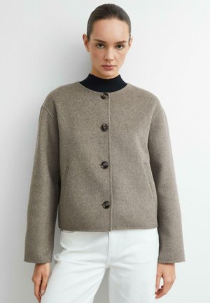 Frau trägt eine taupefarbene, geknöpfte Wolljacke über einem schwarzen Top und weißen Hosen, steht vor einem schlichten weißen Hintergrund.