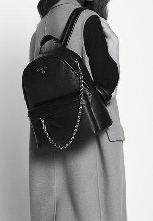 Femme portant un manteau sans manches et un haut noir, avec un sac à dos en cuir noir à détail chaîne porté sur une épaule.