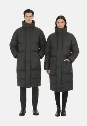 Homme et femme portant des doudounes longues noires à col haut, debout droit avec les mains dans les poches sur un fond blanc.