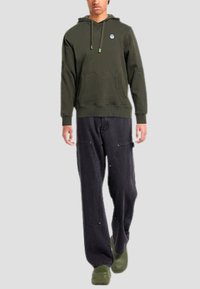 Felpa verde scuro con tasca frontale e coulisse regolabili; abbinata a pantaloni neri larghi e zoccoli verdi. Design semplice e casual.