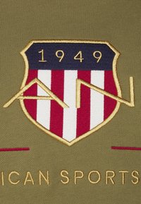 Tela caqui con un logo de escudo bordado que presenta franjas rojas y blancas, azul en la parte superior, el año "1949" y acentos dorados.