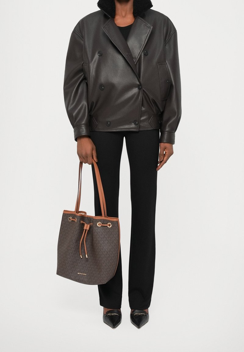 Personne portant une veste en cuir noire et un pantalon noir, tenant un sac fourre-tout en cuir marron avec de longues poignées et une fermeture à cordon.