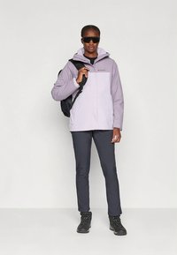 Ljus lavendelfärgad jacka med huva och en logotyp på bröstet, kombinerad med svarta byxor och robusta svarta stövlar. Modellen håller en ryggsäck.