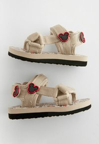 Sandales pour enfants beiges avec brides réglables, semelles noires et icônes de tête de souris noire bordée de rouge sur les brides, avec semelles intérieures à motifs.