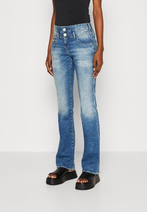 Personne portant un jean bleu délavé à jambes droites avec une taille à double bouton et des sandales noires à plateforme et orteils ouverts, debout sur un sol blanc.