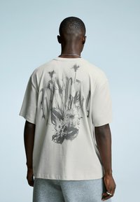Mężczyzna stojący tyłem, ubrany w jasnobeżowy t-shirt z ciemnym kwiatowym i abstrakcyjnym wzorem, w połączeniu z szarymi szortami.