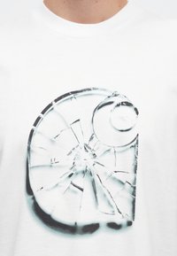 Biele bavlnené tričko s čierno-bielou grafikou prasknutého kruhového dizajnu zobrazujúceho úlomky skla a hladký, zaoblený doplnok.