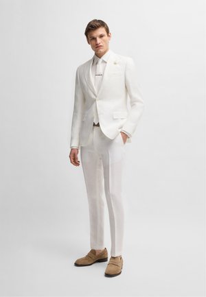 Completo in lino bianco con giacca su misura con rever a ragge e pantaloni abbinati. Scarpe in suede marrone completano l'insieme.