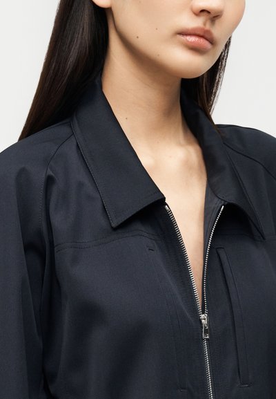 Μαύρο μπουφάν με φερμουάρ που κλείνει, με μυτερό γιακά, υφή υφάσματος και ορατές λεπτομέρειες ραφών. Το φερμουάρ έχει ασημί απόχρωση.