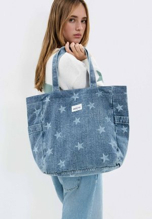 Jeune femme en pull blanc tenant un grand sac cabas en denim bleu avec un motif étoilé et une étiquette de marque visible sur fond uni.