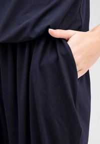 Mörkblå plagg med mjukt tyg, avslappnad passform och sidoficka. Texturen verkar jämn, med lätt drapering runt midjeområdet.