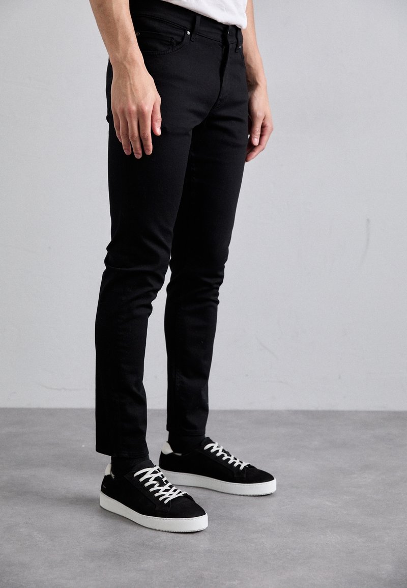 Černé slim-fit džíny s texturovanou tkaninou a předními kapsami, doplněné černými semižovými teniskami s bílou podrážkou a tkaničkami.