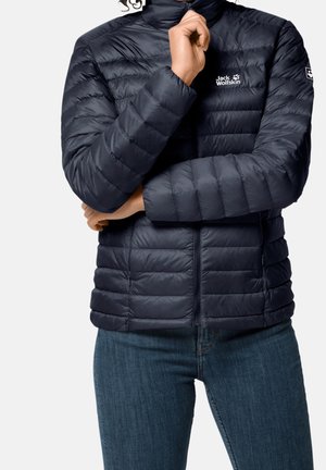 Person wearing a navy blue quilted Jack Wolfskin jacket with zipped front and blue jeans, hand near chin, light gray background.