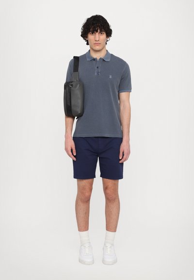 Polo din bumbac gri cu guler, pantaloni scurți bleumarin și o geantă de umăr neagră. Modelul poartă adidași albi și șosete până la gleznă.