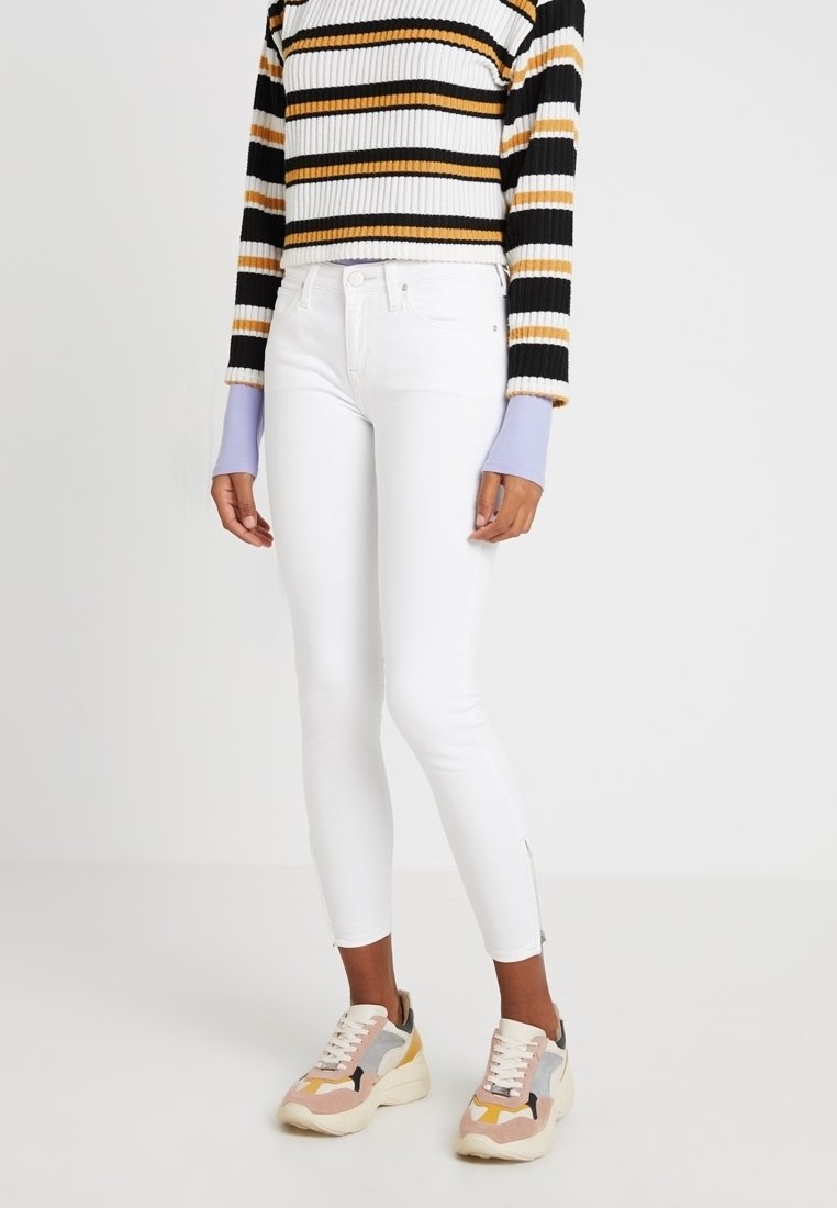 Jeans skinny blancos con cremallera en los tobillos, combinados con un suéter corto a rayas en blanco y negro y zapatillas deportivas gruesas multicolores.