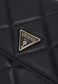 Tikkikuosinen musta keinonahkainen laukku, jossa on kultainen kolmionmuotoinen logomerkki ja tikattu yksityiskohta. Sileä pinta ja kiiltävä viimeistely.