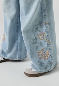 Světle modré džínové kalhoty s širokými nohavicemi a květinovou výšivkou v růžové a žluté barvě, s jemnou texturou a mírně třepenými lemy.