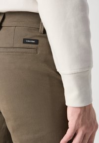 Olivengrønne bukser lavet af bomuldsblanding, med en baglomme og et sort Calvin Klein-logo tag, samt en cremet sweaterærme synlig.