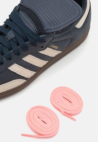 Marineblaue Ledersneaker mit beigen Wildlederstreifen, ausgestattet mit schwarzen Schnürsenkeln und einer strukturierten Zehenkappe. Enthält zwei pinkfarbene flache Schnürsenkel.