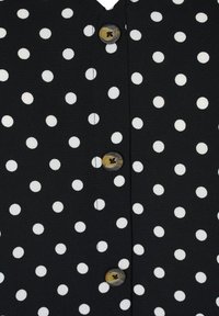 Svart blus med vita prickar, med en V-ringning och fem-knappsstängning med bruna marmorlika knappar. Mjuk, texturerad tyg.