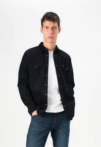 Camisa preta com botões, com dois bolsos no peito, textura lisa e botões de pressão prateados, usada sobre uma camiseta branca e jeans azuis.