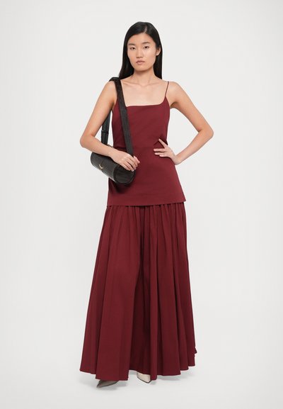 Burgundy two-piece outfit featuring a fitted top with thin straps and a flowy, pleated maxi skirt. Accessories include a black cylindrical bag.