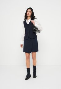 Ναυτικό γιλέκο με κουμπιά, λευκό πουκάμισο και ταιριαστή φούστα. Μαύρη τσάντα και υποδήματα, υφασμένες σκουρόχρωμες κάλτσες. Μινιμαλιστικός σχεδιασμός με καθαρές γραμμές.