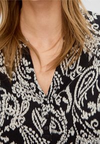 Blusa negra con estampado que presenta un escote en V, mangas cortas y cuatro botones negros en la parte delantera. La textura parece suave con un diseño fluido.