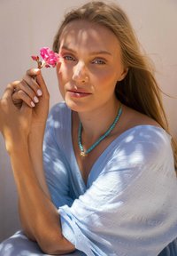 Ljust blått strukturerat tyg draperat över axlarna, med ett turkos pärlhalsband. En hand håller en rosa blomma nära ansiktet. Naturligt ljus.