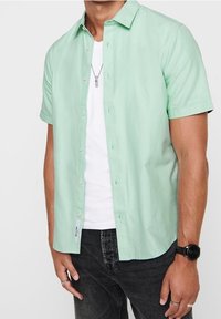 Uomo con camicia a maniche corte verde chiaro button-down sopra una maglietta bianca, jeans neri, orologio da polso, anelli e collana con pendente.