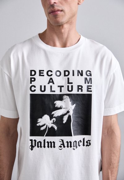 T-shirt en coton blanc avec un design noir représentant des palmiers et le texte "DÉCODER LA CULTURE DES PALMIERS" et "Palm Angels" en lettres majuscules.