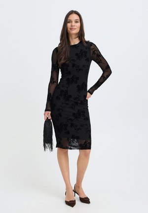 Femme portant une robe fleurie noire transparente, tenant un sac à main noir à franges, debout avec une main sur la hanche et portant des talons pointus foncés.
