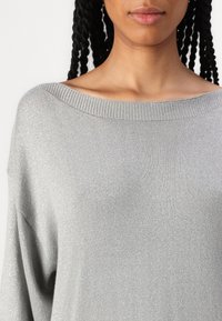 Suéter cinza tricotado com um decote largo em barco, textura suave e brilho subtil. Apresenta um corte descontraído e ombros caídos.