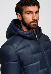 Giubbotto imbottito navy con cappuccio, caratterizzato da un design trapuntato, chiusura con zip e superficie liscia. Dettagli regolabili per migliorare la funzionalità.