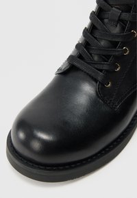 Bottine en cuir noir à bout arrondi, surface texturée et œillets dorés pour les lacets. Détails cousus le long de la semelle.