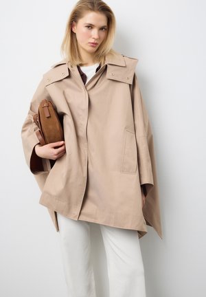 Blondine draagt een beige oversized jas en witte broek, met een bruine leren handtas in de hand, staand tegen een lichtgrijze achtergrond.