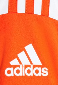 Oranžová látka s bílým logem Adidas. Design obsahuje tři bílé pruhy nahoře, hladkou texturu a výrazné značení.
