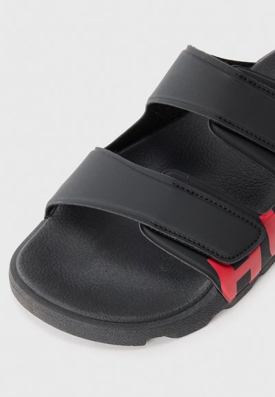 Sandale noire à semelle glissante avec deux larges brides à fermeture Velcro, semelle intérieure texturée et détail du logo rouge sur le côté.