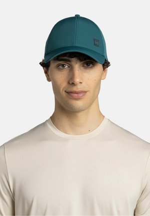 Junger Mann trägt eine türkisfarbene Baseballkappe und ein beiges T-Shirt mit Rundhalsausschnitt, schaut mit neutralem Ausdruck nach vorne vor einem einfarbigen Hintergrund.